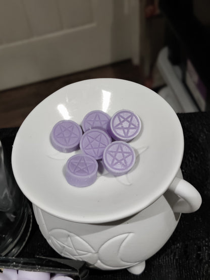 Six lilac purple wax melts with pentagram symbols, arranged on the rim of a white cauldron shaped wax burner.