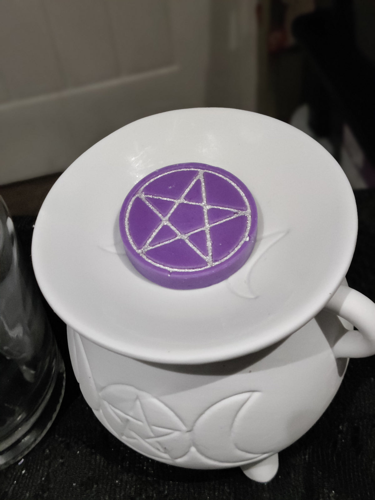 A purple circular wax melt with a silver pentagram design, resting on the rim of a white ceramic cauldron with moon and star embossing.