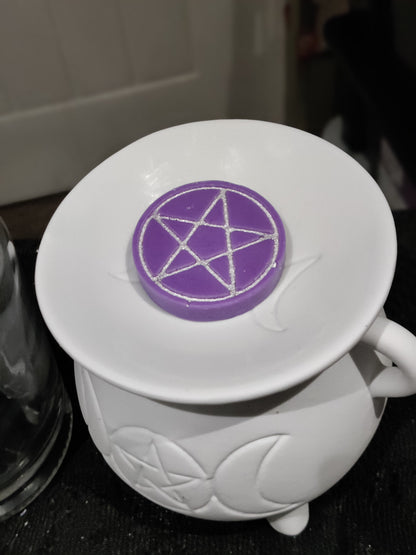 A purple circular wax melt with a silver pentagram design, resting on the rim of a white ceramic cauldron with moon and star embossing.