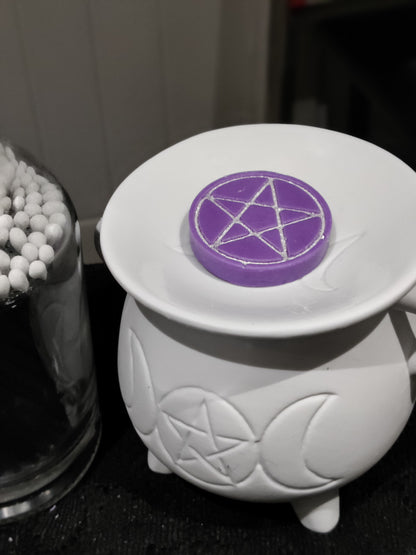 A white ceramic triple moon cauldron-shaped warmer with a purple wax melt featuring a silver pentagram, resting on its dish.