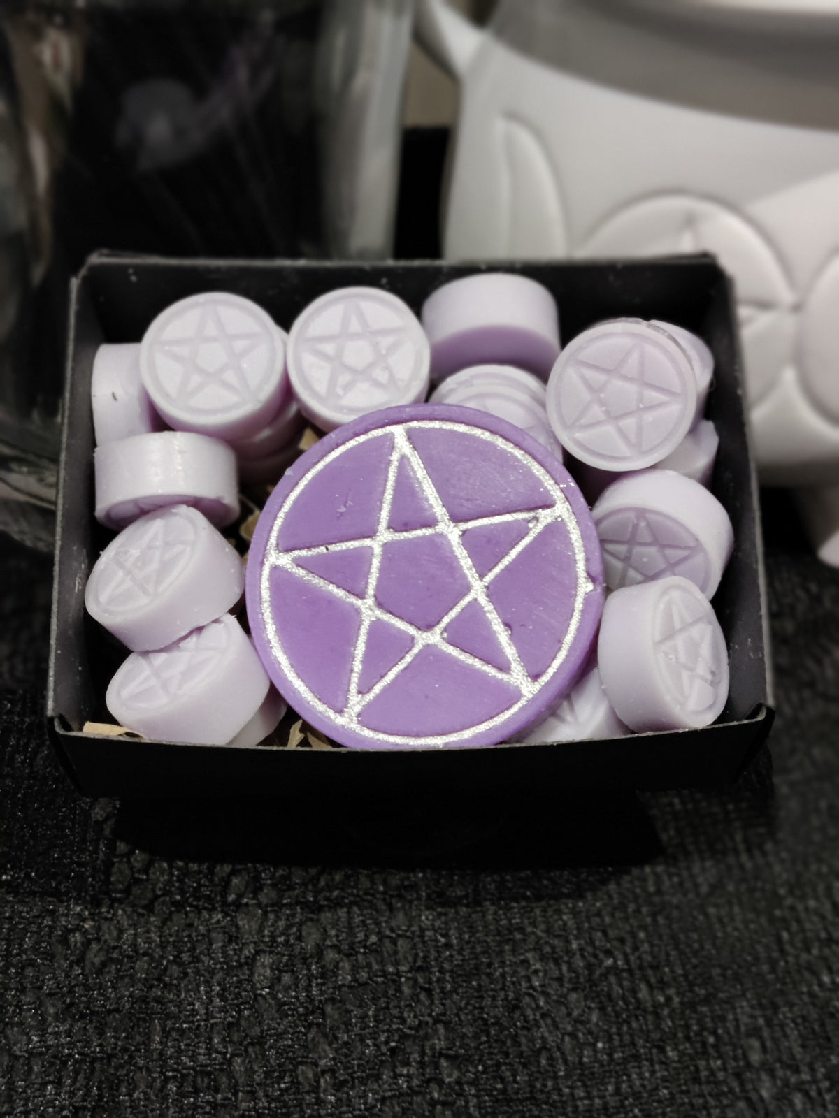 A purple circular wax melt with a silver pentagram design, displayed in a black box alongside several smaller lilac wax melts stamped with pentagrams.