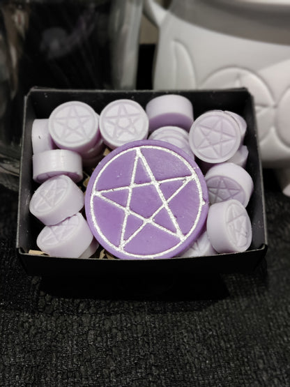 A purple circular wax melt with a silver pentagram design, displayed in a black box alongside several smaller lilac wax melts stamped with pentagrams.