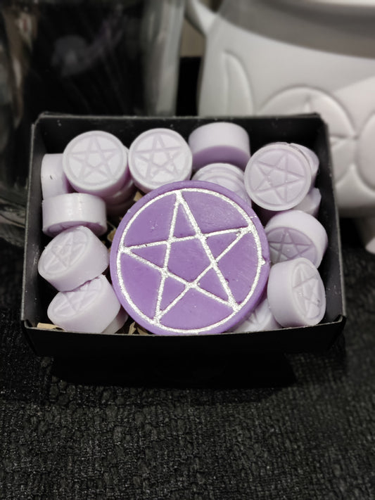 A purple circular wax melt with a silver pentagram design, displayed in a black box alongside several smaller lilac wax melts stamped with pentagrams.
