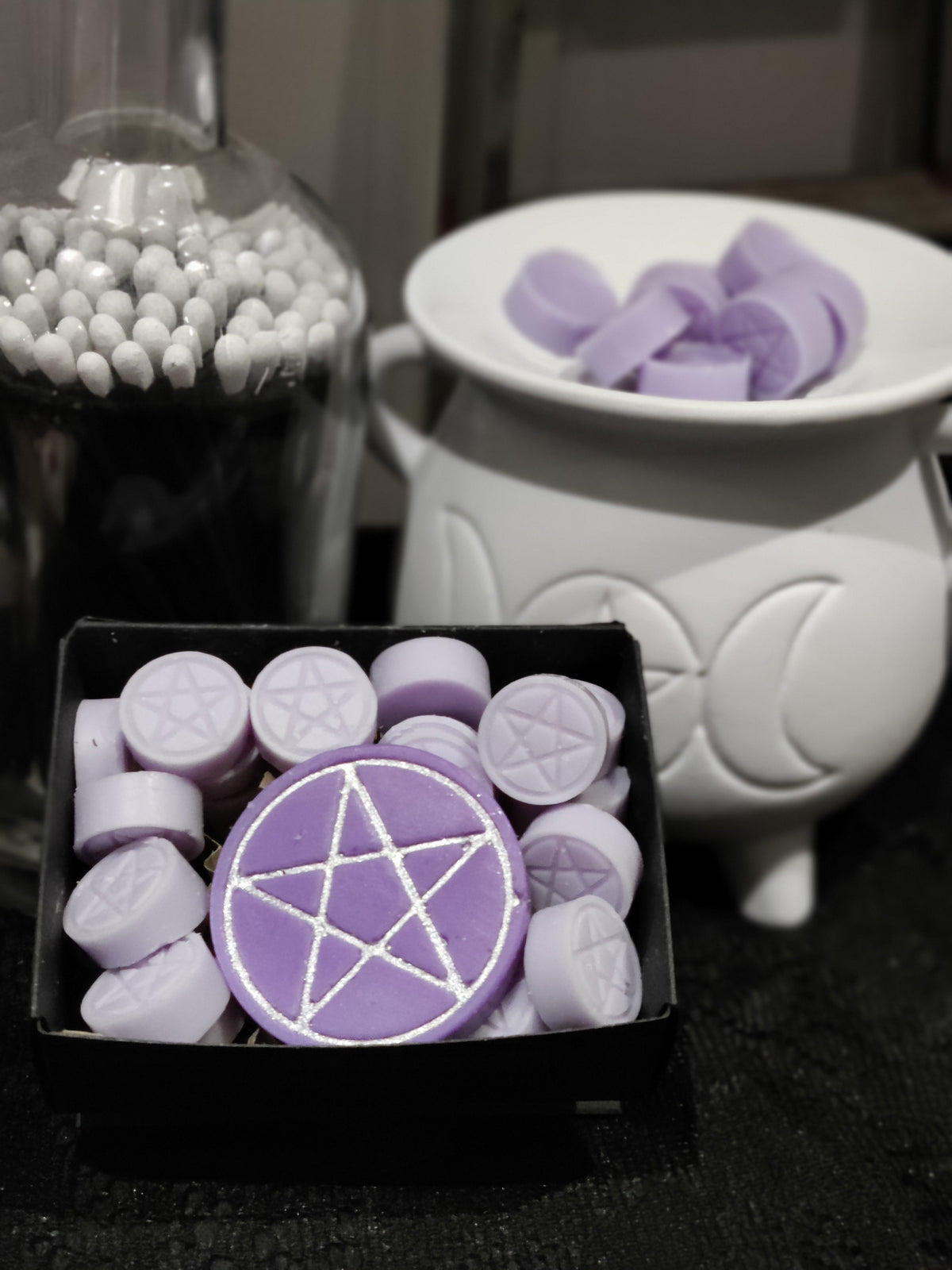 Purple wax melts with silver pentagram designs, shown in a black tray alongside a white cauldron-shaped warmer and a glass jar of long black matches