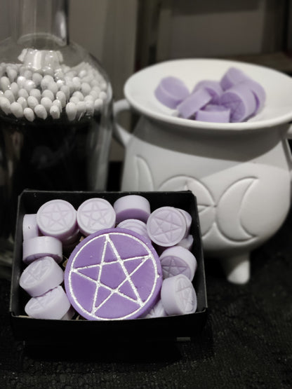 Purple wax melts with silver pentagram designs, shown in a black tray alongside a white cauldron-shaped warmer and a glass jar of long black matches