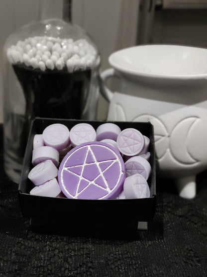 Purple wax melts with silver pentagram designs, shown in a black tray alongside a white cauldron-shaped warmer and a glass jar of long black matches